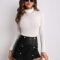 ujer con short falda de cuero sintético negro, top blanco de cuello alto y accesorios, posando contra una pared blanca.